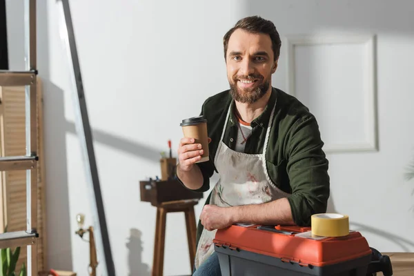 Cheerful carpenter in apron holding takeaway coffee near toolbox in workshop — Foto stock