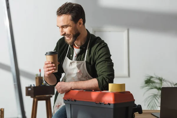 Allegro falegname che guarda il caffè per avvicinarsi al computer portatile e alla cassetta degli attrezzi in officina — Foto stock
