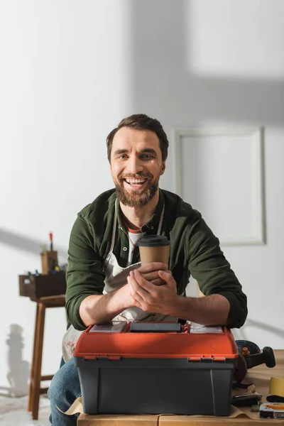 Улыбающийся плотник в фартуке держит кофе, чтобы подойти к ящику с инструментами в мастерской — стоковое фото
