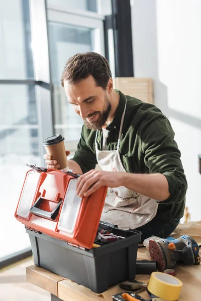 Happy carpenter in apron holding takeaway coffee and opening toolbox in workshop — Foto stock