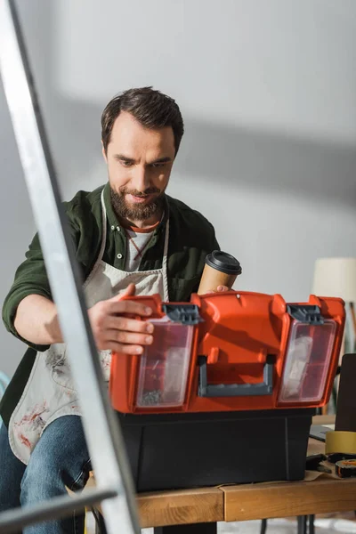 Bearded carpenter holding paper cup and opening toolbox in workshop — Stock Photo