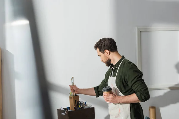 Side view of carpenter holding coffee to go and taking paintbrushes in workshop — Foto stock