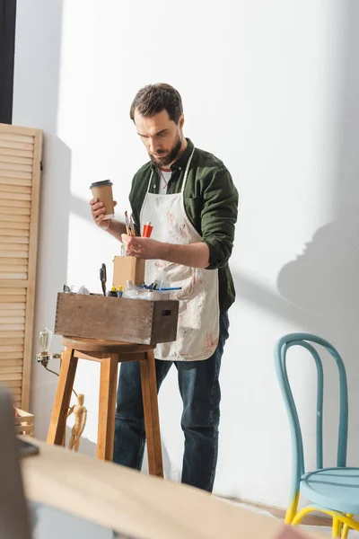 Ремесленник в фартуке держит кисти и кофе, чтобы пойти в мастерскую — стоковое фото
