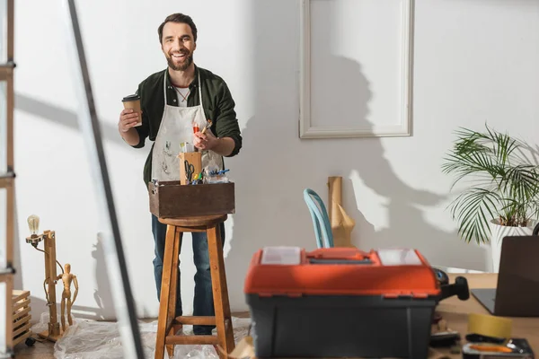 Artigiano sorridente che tiene il caffè per andare e pennelli in officina — Foto stock