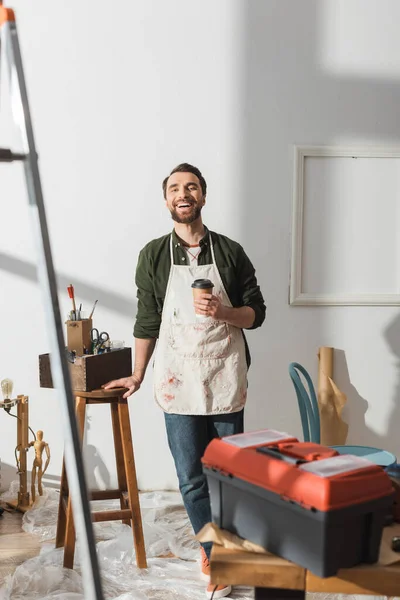 Positive carpenter holding coffee to go and looking at camera in workshop — Foto stock