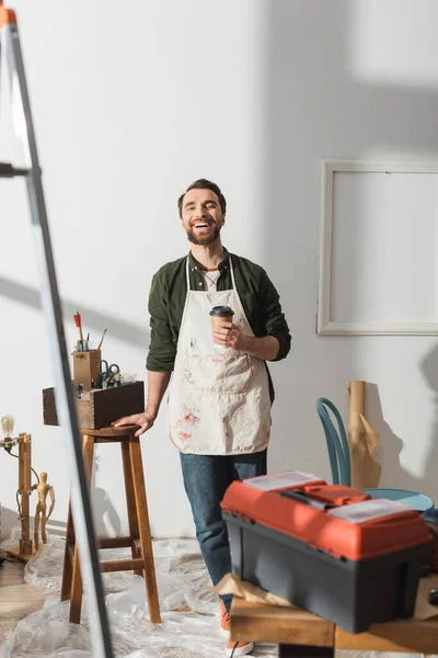 Positive carpenter holding coffee to go and looking at camera in workshop — Stock Photo