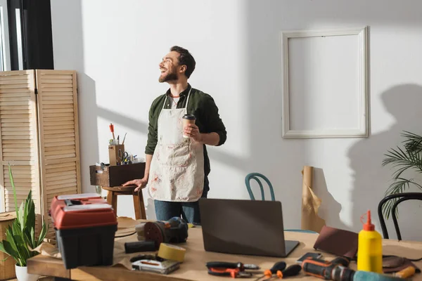 Alegre carpintero en delantal sosteniendo café para acercarse a herramientas en taller - foto de stock
