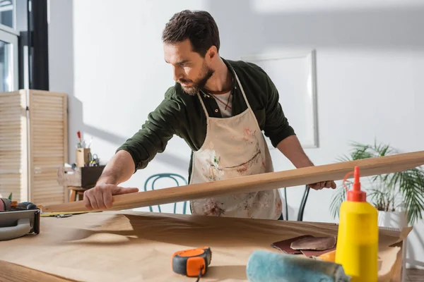 Carpenter in dirty apron putting wooden board on table near ruler and sandpaper — Stock Photo