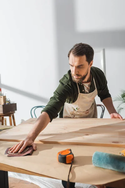 Menuisier barbu prenant du papier de verre tout en travaillant avec une planche en bois dans l'atelier — Photo de stock