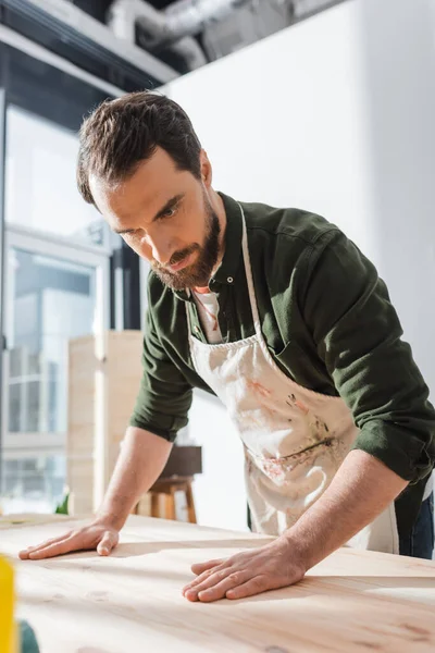 Bearded craftsman in apron touching wooden board in workshop — Stock Photo