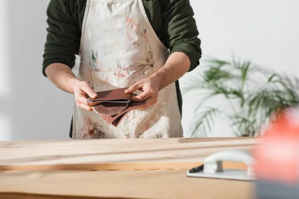 Обрезанный вид рабочего в фартуке с наждачной бумагой рядом с деревянной доской — стоковое фото