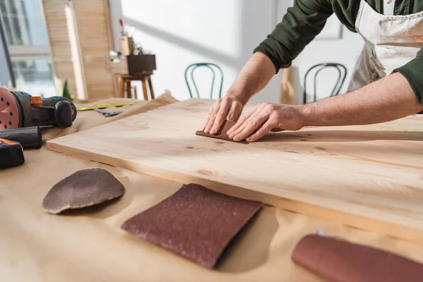 Cropped view of repairman sanding wooden board near sandpaper in workshop — Stock Photo