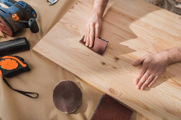 Верхний вид плотника, шлифующего деревянную доску рядом с инструментами на столе — стоковое фото