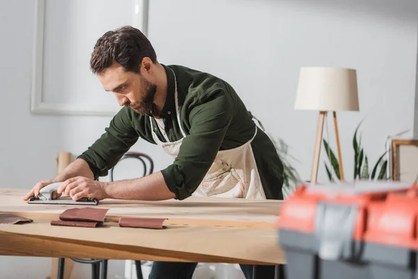 Repairman in apron sanding wooden plank near blurred toolbox in workshop - foto de stock
