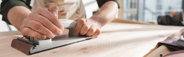 Cropped view of man in blurred apron sanding wooden board in workshop, banner - foto de stock