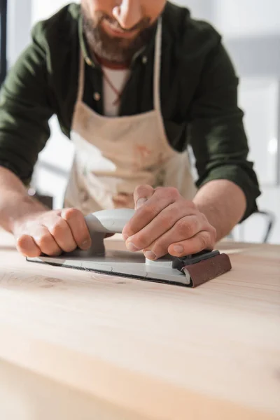 Cropped view of blurred bearded restorer sanding wooden board in workshop — Stock Photo
