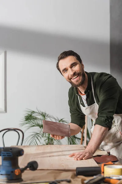 Smiling carpenter in apron holding sandpaper near board and looking at camera in workshop — Stock Photo