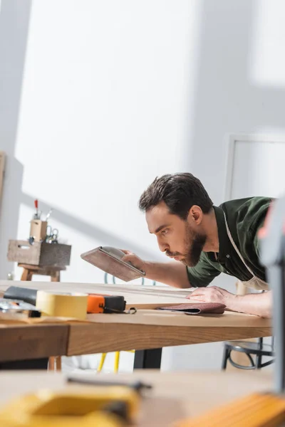 Brünetten Tischler hält Schleifpapier und bläst auf Holzbrett in Werkstatt — Stockfoto