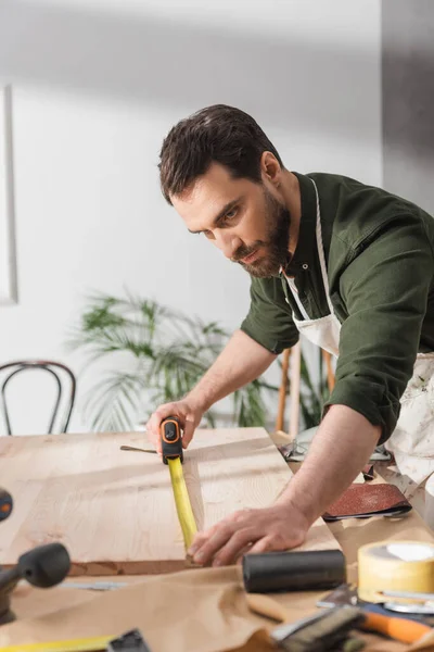 Restorer in apron measuring wooden board with ruler near tools in workshop — Foto stock