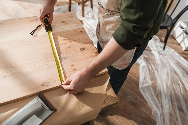 Высокоугловой вид плотника в фартуке, измеряющий деревянную доску в мастерской — стоковое фото