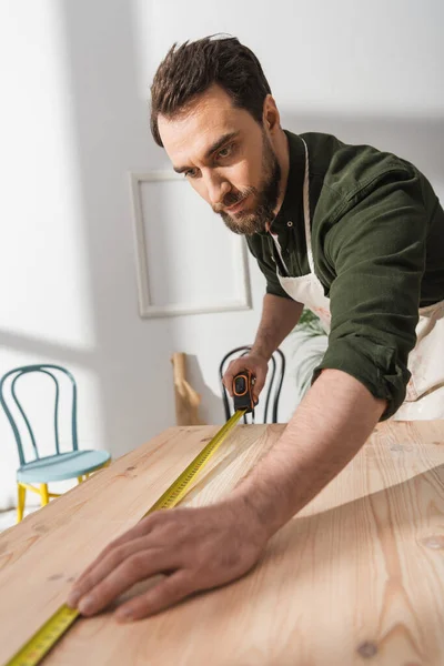 Bearded carpenter in apron measuring wooden board in workshop — Foto stock