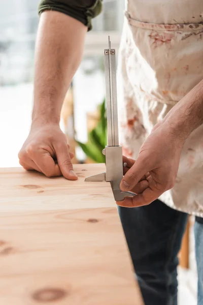 Cropped view of craftsman in dirty apron measuring wooden board with calipers in workshop — Photo de stock