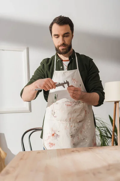 Bearded restorer in apron looking at calipers near blurred wooden board — Stock Photo
