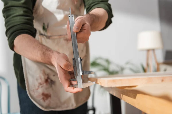 Cropped view of blurred restorer in apron holding calipers near wooden board — Stock Photo