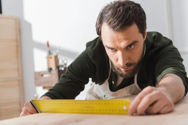 Focused carpenter looking at square tool on blurred wooden board - foto de stock