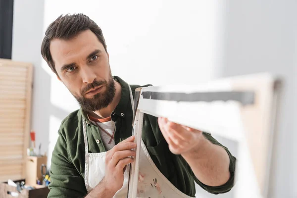 Bearded repairman in apron looking at blurred picture wooden frame in workshop — Stock Photo