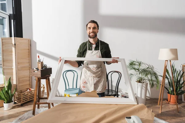 Positiver Schreiner blickt in Kamera, während er Holzgestell auf Tisch in Werkstatt hält — Stockfoto