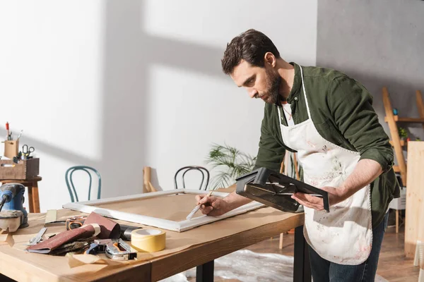 Bearded restorer in apron painting on picture frame in workshop — Stock Photo