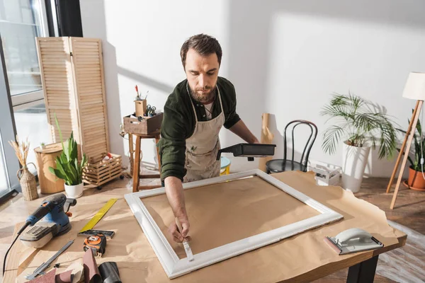 Peinture restaurateur barbu sur cadre photo en bois près des outils en atelier — Photo de stock