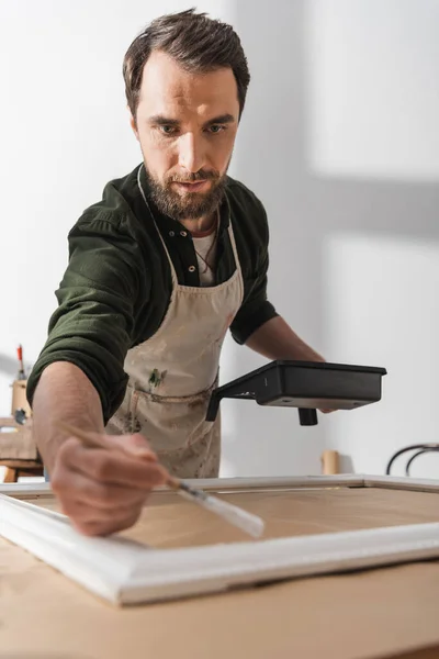 Restaurateur concentré dans le tablier tenant le pinceau près du cadre flou dans l'atelier — Photo de stock