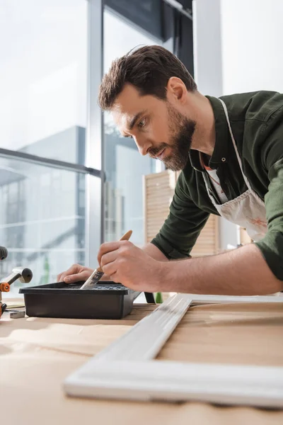 Bearded restorer holding paintbrush near paint and blurred wooden picture frame in workshop — Stock Photo