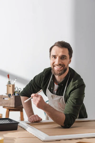 Cheerful restorer in apron holding paintbrush and looking at camera near wooden frame in workshop — Stock Photo
