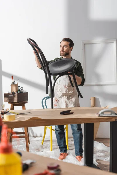 Bearded restorer holding chair near working table in workshop — Foto stock
