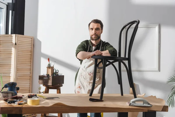 Bearded restorer in apron looking at camera near wooden chair in workshop - foto de stock