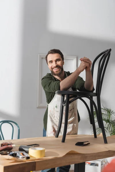 Positive restorer in dirty apron standing near chair on table in workshop — Foto stock