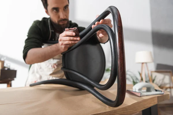 Размытый ремесленник в фартуке шлифовка черного деревянного кресла в мастерской — стоковое фото