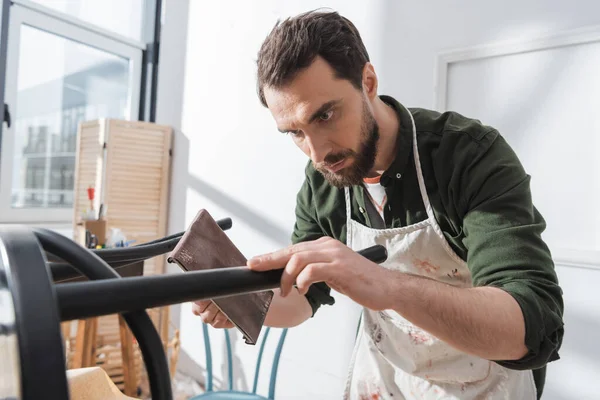 Restorer in dirty apron sanding paint on wooden chair in workshop — Foto stock