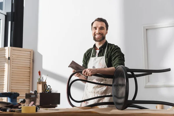 Smiling restorer in apron holding sandpaper near chair and looking at camera in workshop — Stock Photo