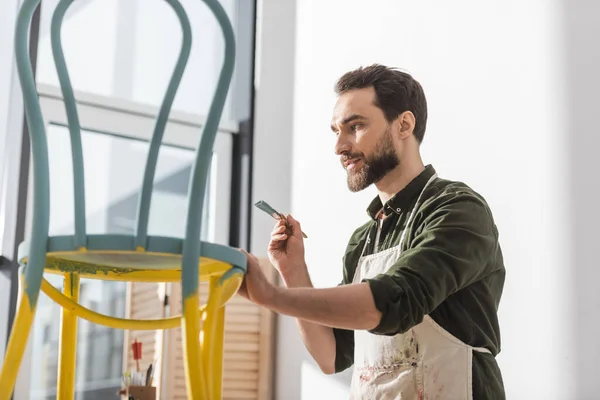 Restauratore barbuto in grembiule con pennello vicino alla sedia in officina — Foto stock