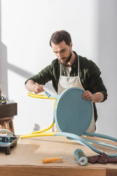 Craftsman in apron repainting chair near roller on table in workshop — Stock Photo