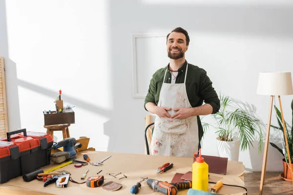 Positive restorer in apron looking at camera near tools on table in workshop — Fotografia de Stock