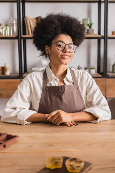 Happy african american woman in eyeglasses and apron sitting near blurred soap bars on table in craft workshop — Stockfoto