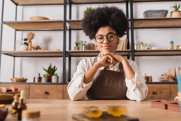 Happy african american woman sitting near blurred handmade soap on table in workshop — Foto stock