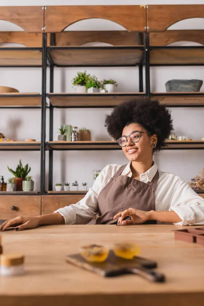 Joyful african american craftswoman in apron and eyeglasses sitting near blurred handmade soap on table in workshop — Photo de stock