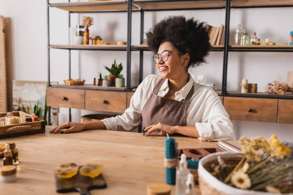 Happy african american craftswoman sitting in workshop near handmade soap and natural ingredients on blurred foreground — Stock Photo