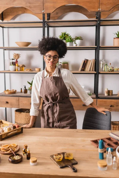 Smiling african american woman in eyeglasses and apron standing near natural ingredients for soap and candle making — Stockfoto